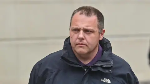 A man with short dark hair stands in front of a grey-coloured wall. He is wearing a navy waterproof jacket and a purple polo shirt. 