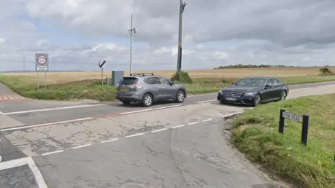 Google Street View image of a crossroads surrounded by farmland with two cars passing in opposite directions