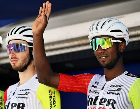 LUC CLAESSEN / GETTY IMAGES Two men dressed in racing lycra, helmets and sunglasses stand side by side. One of them waves.
