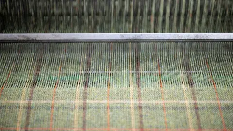 A green tweed being woven on a loom. There is pattern of brown, orange and yellow through the fabric