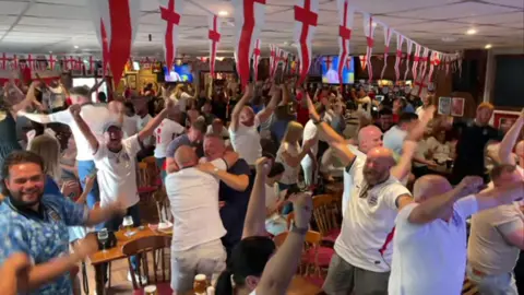 A crowd of people in mainly England shirts in a pub or club celebrating a win with arms aloft and in some cases hugging each other
