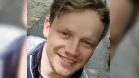 Hampshire and Isle of Wight Constabulary Craig Laurie smiles in a close up photo. He has wavy brown hair and a short brown beard, and is wearing a zip up jacket. 