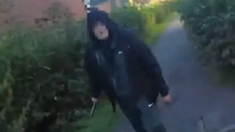 A man in a black hooded top and a tracksuit. He is standing on a pavement with bushes either side of him, in a residential area. He is holding a large knife in his right hand. His face is blurred to obscure his identity.