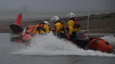 RNLI Three RNLI crew onboard an orange inshore lifeboat. They all wearing red life jackets, white helmets and yellow jackets. Water is spraying up the sides of the boat as it travels through the water in foggy conditions.