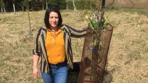 Natalie Paul Natalie has shoulder length brown hair and is wearing a yellow and black stripy top. She is leaning against the memorial tree.