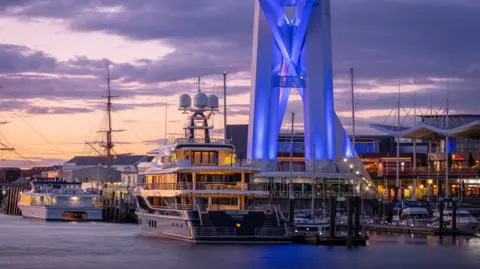 View from feet today A luxury yacht is moored in Portsmouth harbour next to the base of the Spinnaker Tower. The tower is lit up blue. Behind you can see the buildings of the Gunwharf centre lit up. And in the background you can see other boats and a large mast against the evening sky.