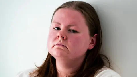 A young woman's head, with her sitting against a wall, with long hair, looking at the camera with an intense expression