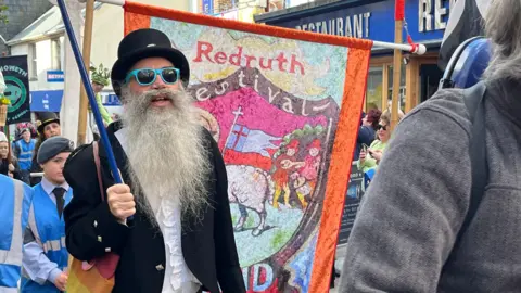 A man with a long grey beard wearing a top hat and blue sunglasses walks in the parade in Redruth. 