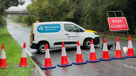 A road closed to oncoming traffic with a line of orange and white cones. Behind the line is a red sign that reads "Road Closed" and a white van with "Milton Keynes City Council" logo on it. 
