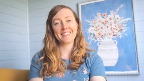 Sian Lawley-Rudd Sian Lawley-Rudd, a women with long blonde-brown hair smiles while sitting on a sofa in front of a painting of flowers. She is smiling and wearing a blue t-shirt with dogs on it.