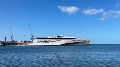 A large, white trimaran ferry with the word Condor emblazoned on the site, along with yellow and pink ribbons, pulls away from an old granite pier on a sunny day.