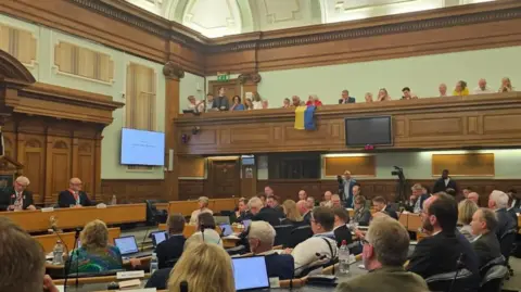 A council chamber full of people with a yellow and blue Ukrainian flag draped over the public gallery balcony