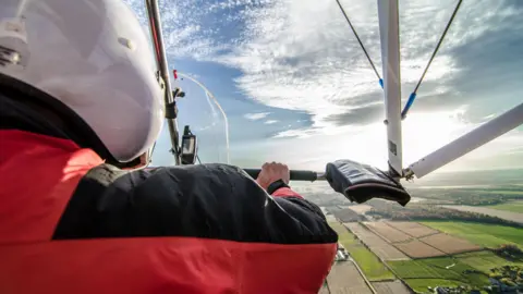 A view from inside a microlight aircraft over the pilot's shoulder to fields below