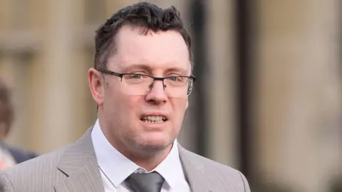 PA Media Image shows man in a light-brown suit with a silver tie and black-rimmed glasses. He has black hair and grey stuble.