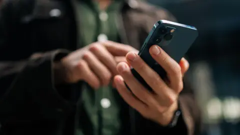 An unrecognisable man's hand holding a smartphone. Only the back is visible. In soft focus behind the hand holding the phone, his other hand typing on the phone, his green shirt, and his black jacket are visible.