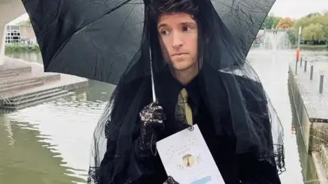 BBC Radio 1 Greg James looking sad dressed in an all black mourning outfit. He's holding a black umbrella and holds an order of service for a service honouring long boi. He's near a body of water.