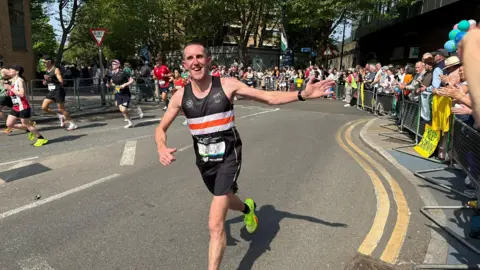A man in a black, white and orange running vest with race number visible runs along a London street on a sunny day, reaching out towards spectators who line metal barriers on the right. Other runners are visible behind him and trees are in full leaf overhead.
