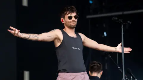 Getty Images Louis Tomlinson performs on stage wearing a black t-shirt with "British Boy" written on it. He is also wearing sunglasses and pale pink trousers. 