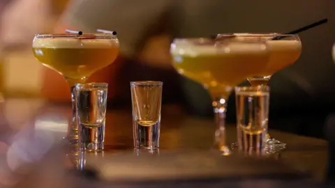 Three cocktail glasses with a yellow frothy liquid inside. There are also three shot glasses placed next to the glasses. 