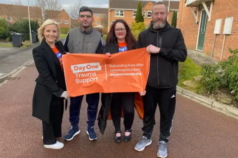 Day One Trauma Support Cat Neill stands with three other people holding up a "Day One Trauma Support" banner. They are on a residential street.