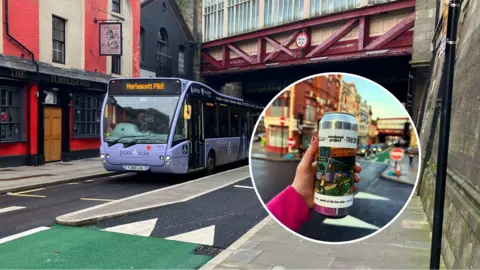 A mauve bus at a bus stop near a railway bridge with an inset photo of a can of beer