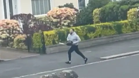 Man running down street with chainsaw. He is wearing a grey hoody and black trousers.