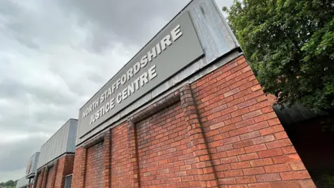A red brick building, viewed from a corner, with metal flashing around the top. There is a grey sign with lettering that reads North Staffordshire Justice Centre.