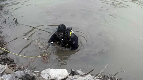 South Wales Police A diver in full diving gear emerges on the shore of a pond full of dark silt