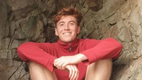 Tomos Sleep who has short light brown hair wearing a red jumper and shorts sat down.