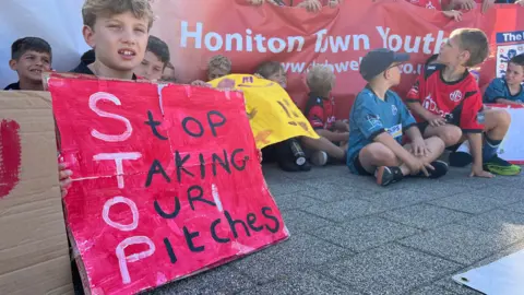 Children sat protesting. They are holding banners and painted signs. One reading "Stop taking our pitches".