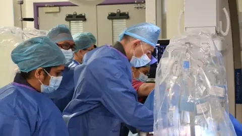 A group of medical professionals in blue scrubs and facemasks. They are all gathered together and looking down at a patient - who is out of shot.