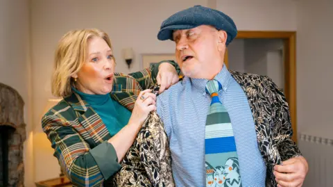 BBC/One Tribe TV Joanna Page is putting a jacket onto a man who is wearing a hat and looking at her.