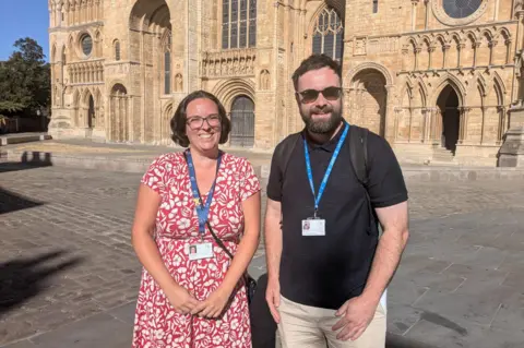 A man and a woman smile as they stand in front of a huge medieval stone cathedral, built in the Gothic style. The woman has brown hair in a bob and wears glasses and a red floral print dress. The man has short brown hair and a full beard and wears sunglasses, a black polo shirt and beige trousers.