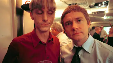 Two men stand side by side, one in a dark red shirt and one in a blue shirt with a dark tie. The man in red is looking sideways at the other, holding what looks like a pint of beer in a plastic cup.