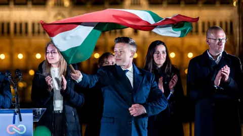 Magyar pictured in the dark holding the flag of Hungary and looking towards left of frame in front of other party members clapping with parliament in background as they celebrate after winning the election.