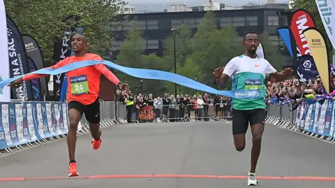 Pacemaker Abay Alemu has a page saying number 24 pinned to his top. Teadese Mamo has a number 25 pinned to his top. They are running through a blue ribbon. People are watching from behind barriers. 