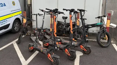 An Garda Síochána Several orange and black e-scooters beside a white An Garda Síochána van