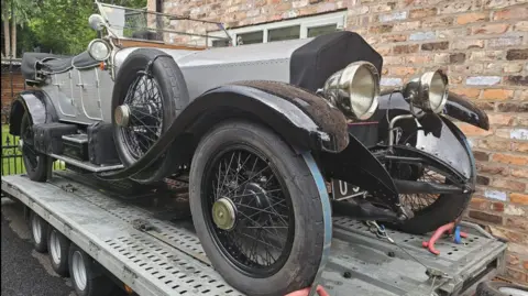A 1920 Rolls Royce Silver Ghost car, pictured on a metal flat-bed trailer. The car is open-top with a grey body and black running board 