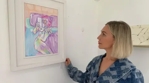 BBC Clíodhna Doherty regarde sa peinture Laura dans son studio d'art à Newry. La peinture est une œuvre d’art figuratif abstrait montrant une femme nue lisant un livre au lit. Clíodhna a un carré blond et porte une robe en jean bleu.