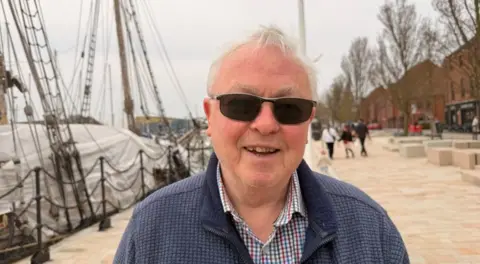 Barry wears a pair of sunglasses and smiles for the camera. A boat and the new path can be seen in the background.
