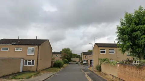 A road leads towards rows of mustard-coloured stone houses, with slanted brown roofs. Lamposts and cars can be seen parked in the background.