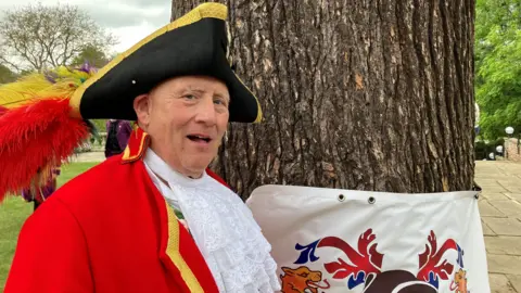 Henry Godfrey-Evans/BBC A man in a classic red town crier's outfit and a black hat, standing next to a tree trunk with a banner tied around it