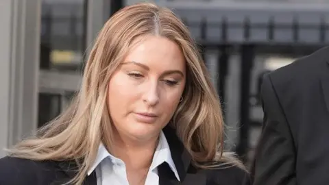 PA Media Nikita Hand has long blond hair and is wearing a dark suit jacket and open-necked shirt. She is looking downwards as she leaves court