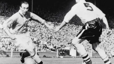 A black and white photo of Sir Stanley Matthews in action on the pitch during the FA Cup final in 1953.