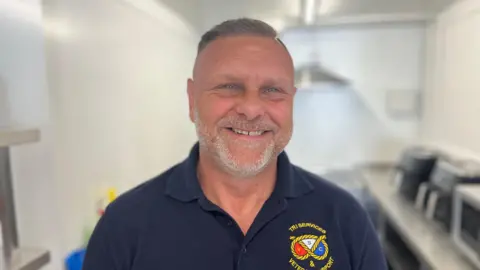 A man in a navy blue shirt with a Tri Services badge on it, smiles while standing in a kitchen.