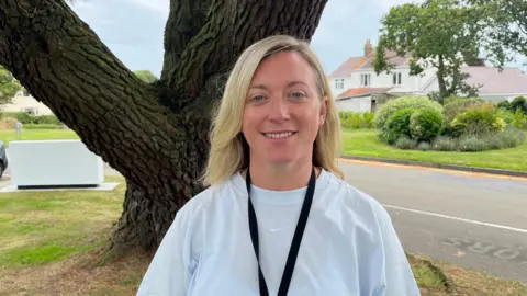 A blonde woman in a white T-shirt stands in front of a tree.
