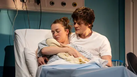 Johan Persson A couple in a hospital bed. Aramayo wears a white t-shirt. Sheehy is in a white and blue hospital gown, and is holding a baby wrapped in a white blanker
