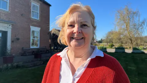 BBC A woman with blonde hair, a white shirt and red cardigan smiles as she stands in front of a building and garden.