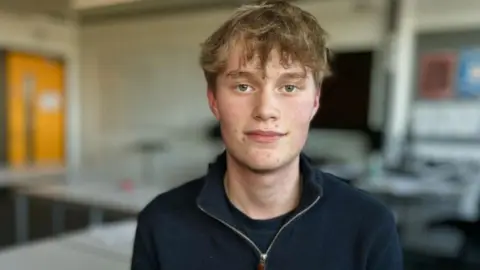 Martin Giles/BBC Will in the classroom in Bury St Edmunds in Suffolk. He has brown hair and is wearing a blue zip-up top over a blue t-shirt.