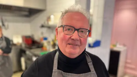 David Savident - A man with white spiky hair looking towards the camera, wearing blue-rimmed glasses. He's wearing a black jumper and a grey apron. 